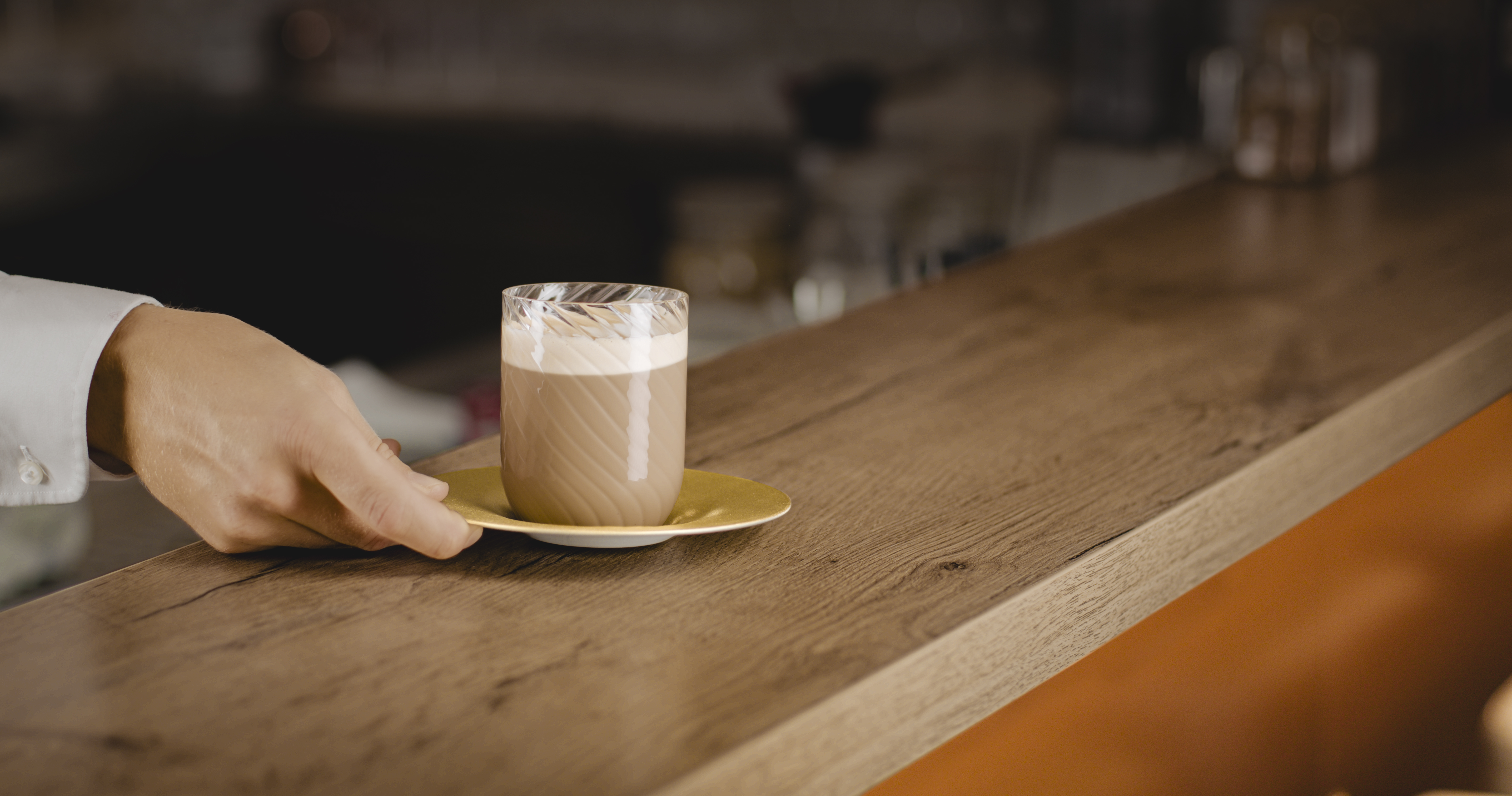 Eine Hand stellt einen Kaffee mit Golduntertasse auf eine Bar