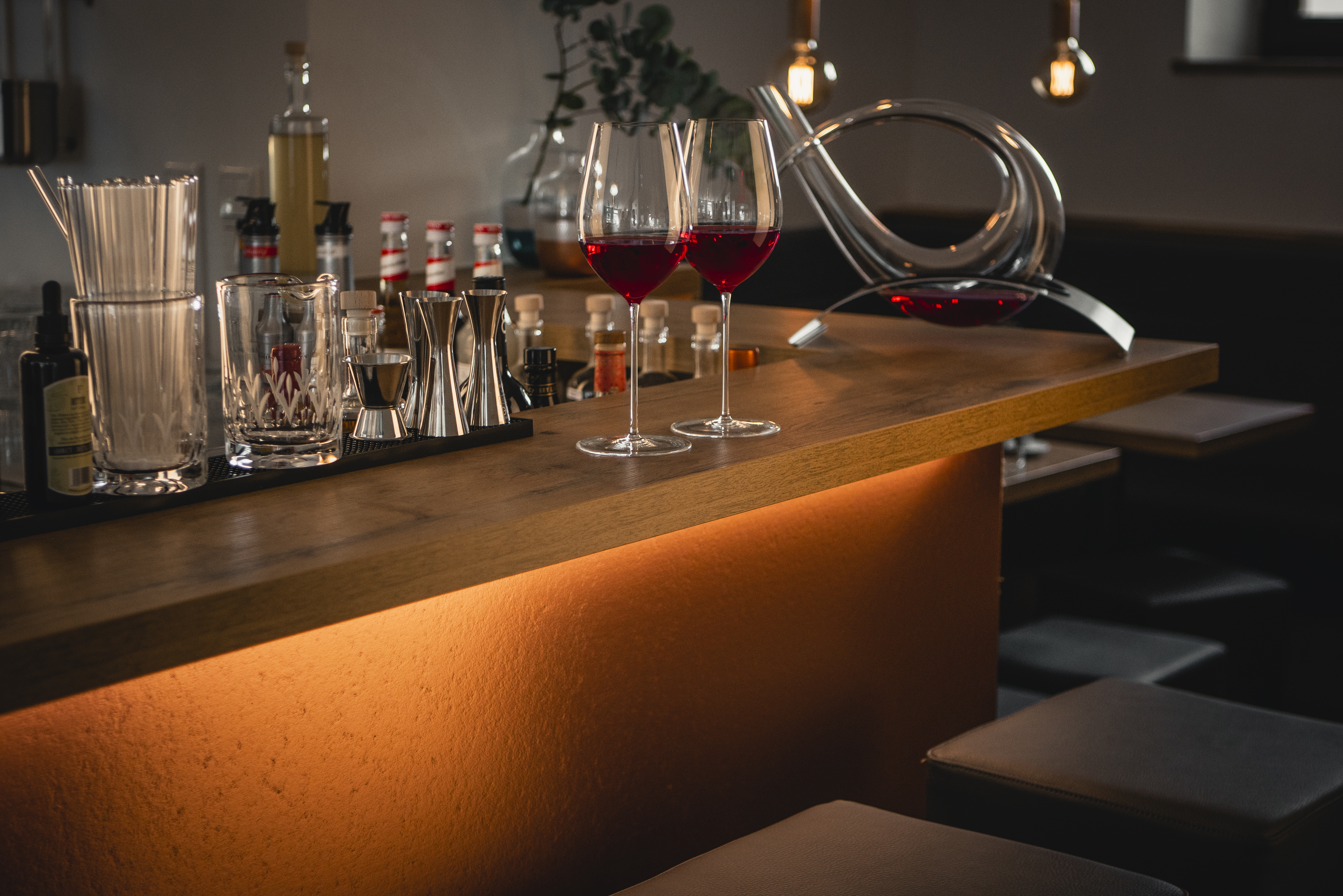 Two filled wine glasses with a decanter on a bar