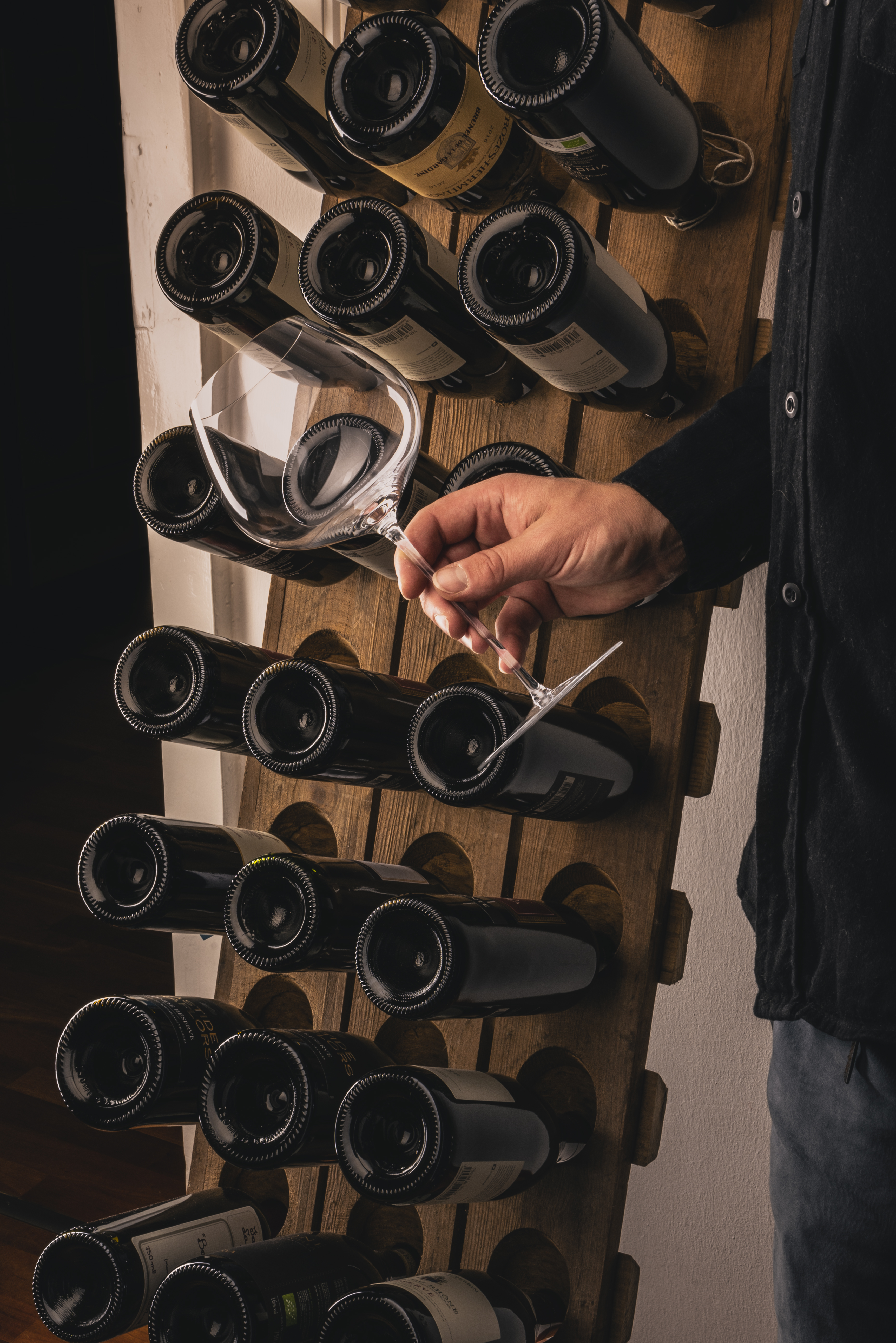 Hand holding a glass in front of a collection of wine bottles