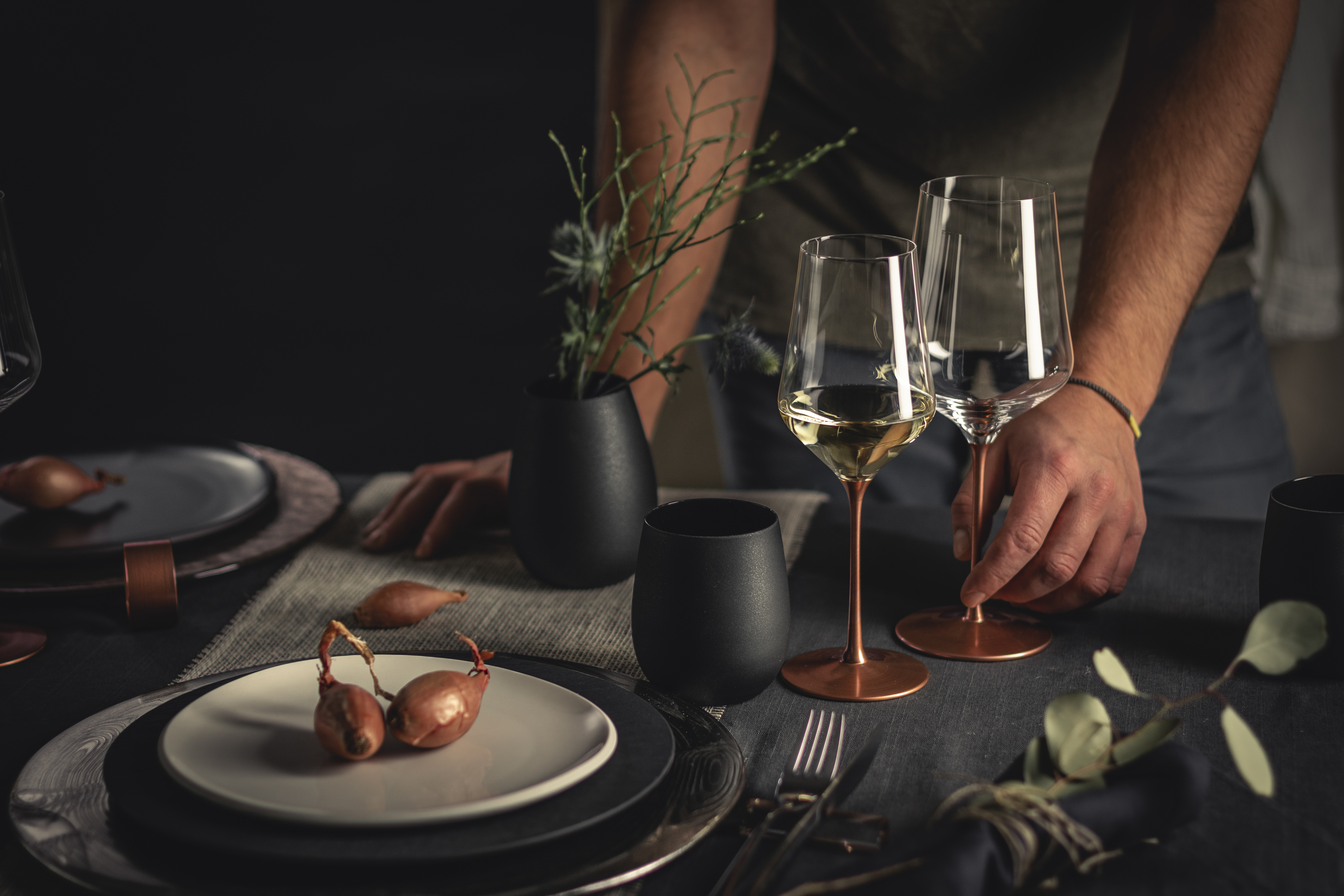 A hand places a second wine glass with copper stem on a beautifully laid table
